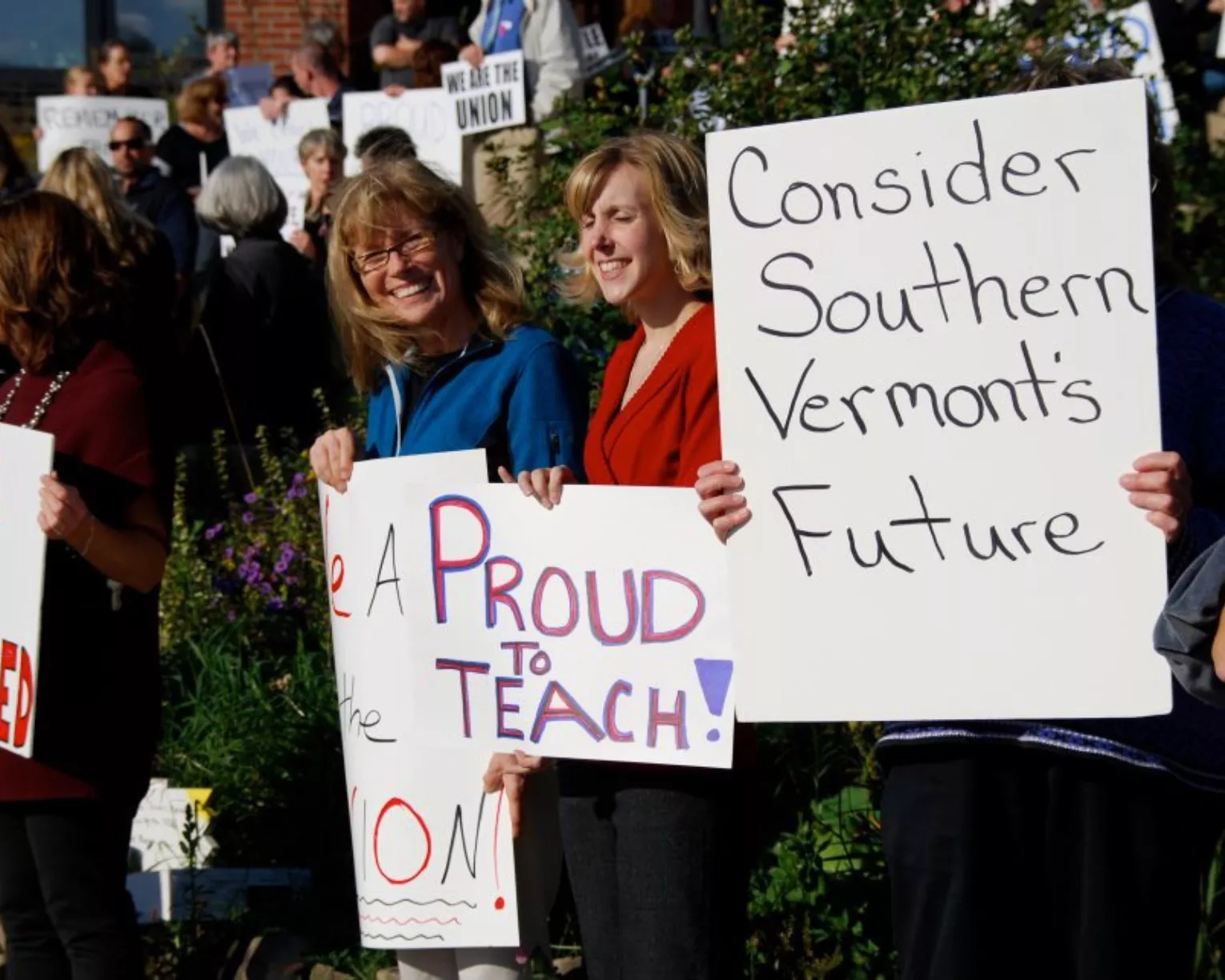 VT teachers stand smiling with signs; one says "Proud to Teach."