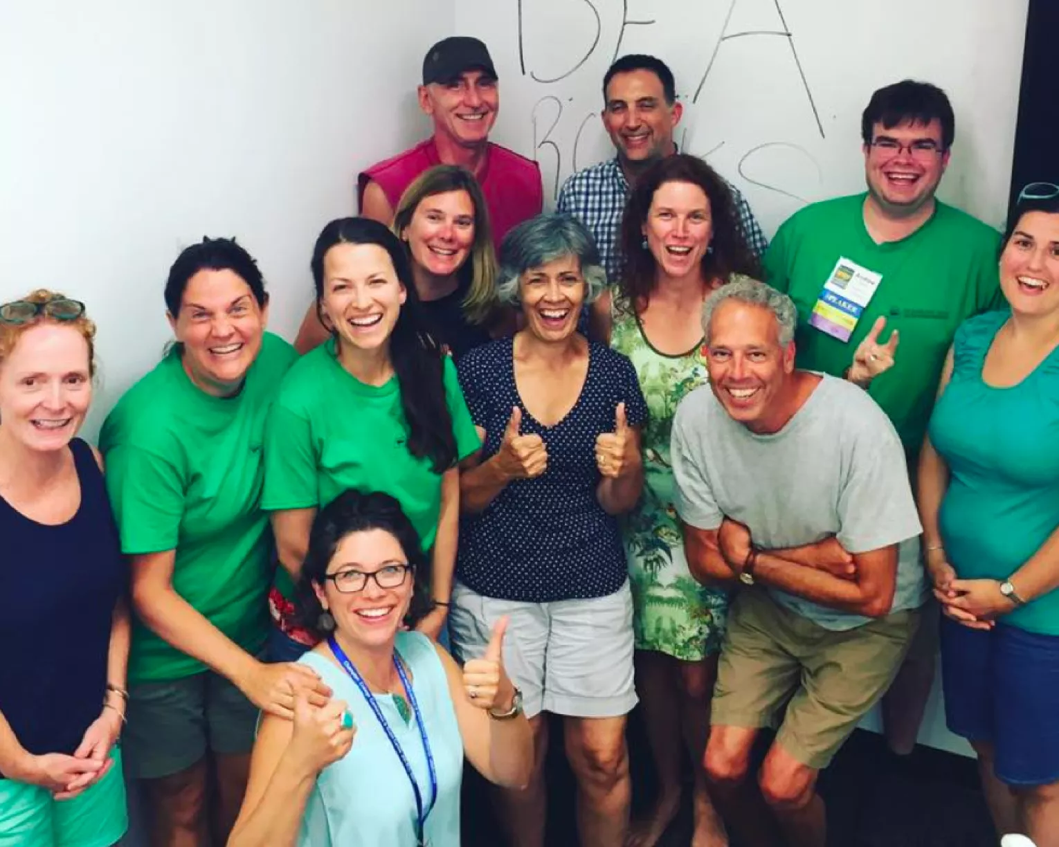 Members stand together in a group in green shirts smiling with thumbs up
