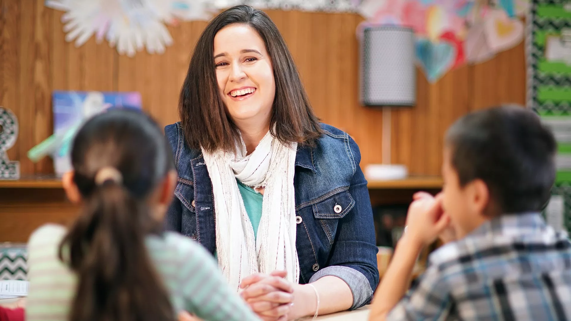 A teacher sits smiling in front of her class. You can see the back of two students' heads.
