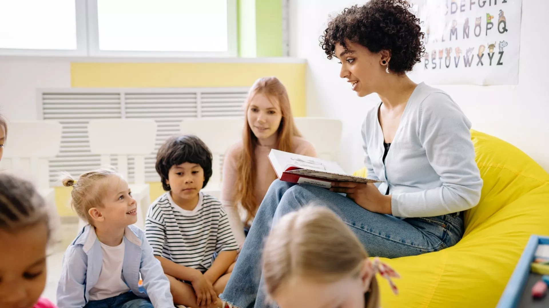 A teacher sits on a yellow chair reading to her kindergarten class.