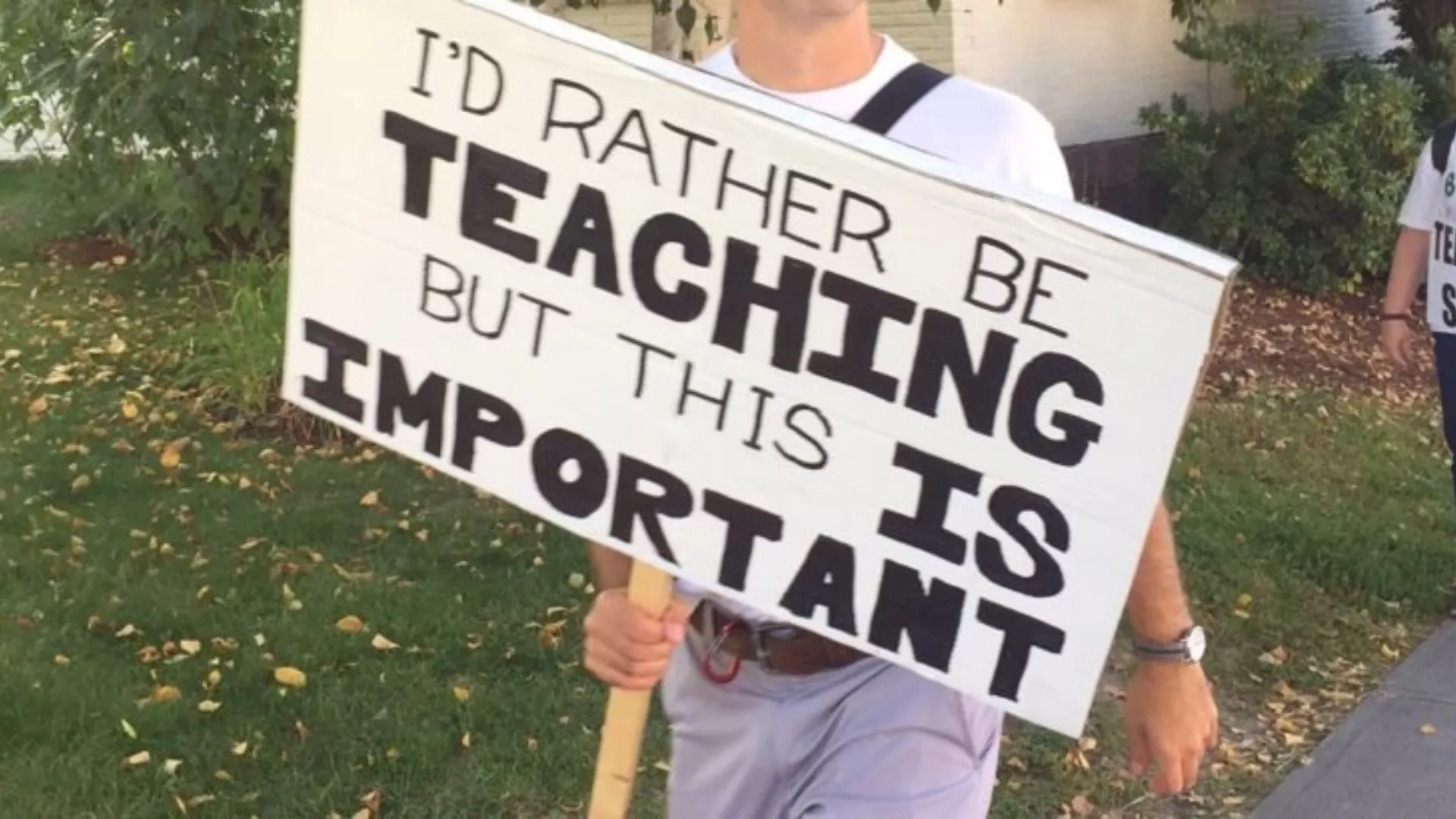 An unidentified teacher whose face is not pictured holds a sign that says, "I'd rather be teaching, but this is important."