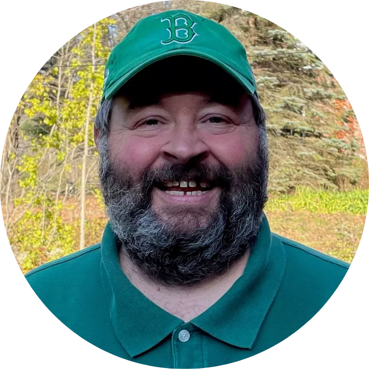James "Jim" Johnson poses for a photo with a big smile in a green hat and shirt. Deciduous and coniferous trees can be seen in the background.